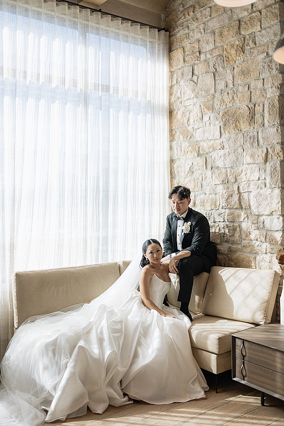 Couple portrait of bride and groom on sofa, her strapless gown and long veil flowing as he wears a black tux by a stone wall window