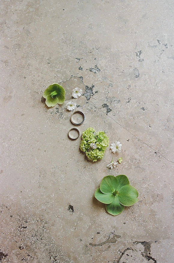 Wedding rings flatlay with a diamond engagement ring and bands nestled beside green hydrangea and small white flowers on stone surface