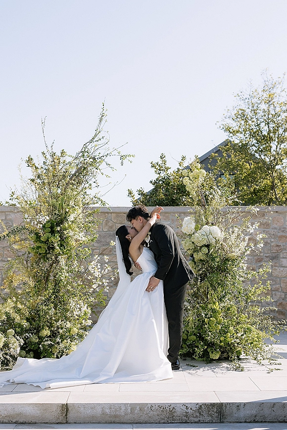 Wedding kiss portrait of bride and groom kissing beneath a greenery and white floral arch, long veil flowing against stone wall and blue sky