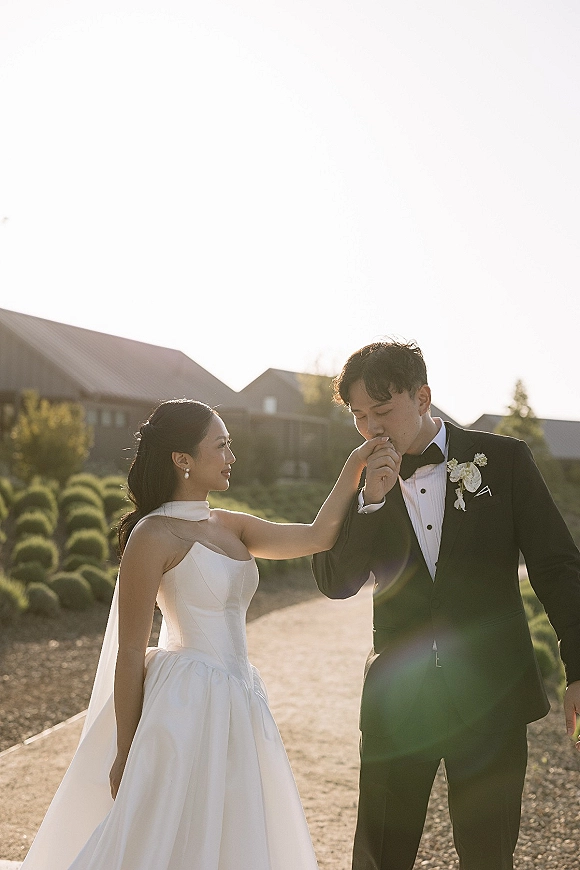 Couple portrait of bride and groom holding hands as he kisses her hand, veil and tuxedo glowing at golden hour on a garden path