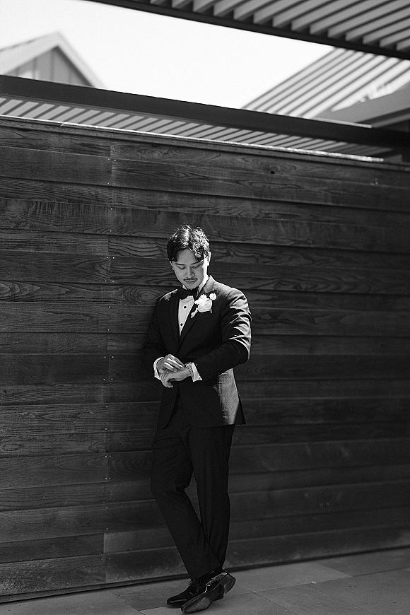 Groom portrait in a black tuxedo adjusting cufflinks, bow tie and boutonniere visible, leaning against a wood wall backdrop