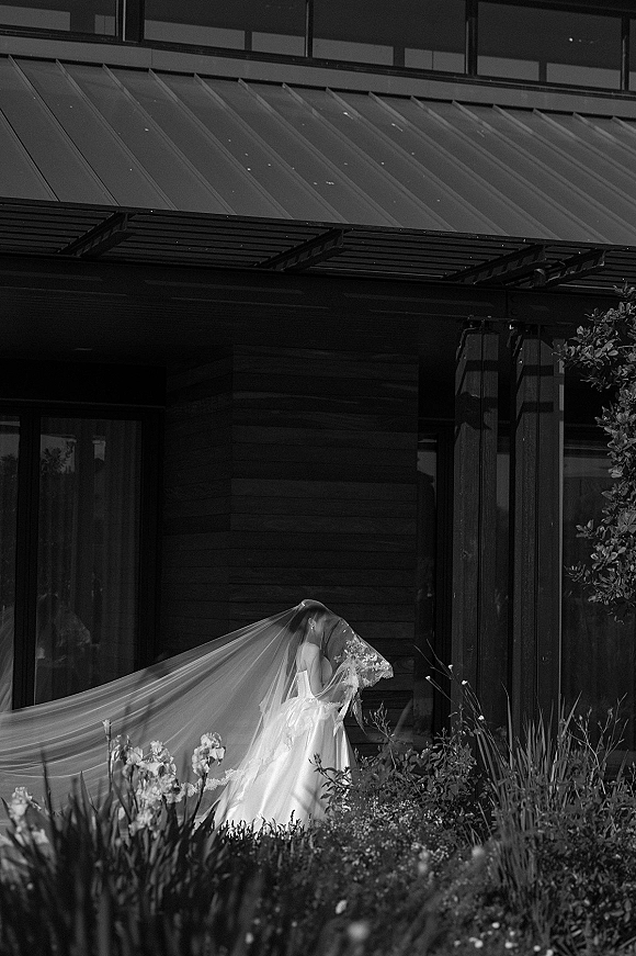 Bridal portrait of a bride with long veil holding a bouquet over her face in a strapless gown outside a modern glass building with garden flowers