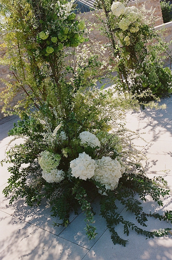 Wedding floral installation of white and green hydrangeas with greenery garland and tall branches against a sunlit stone wall patio