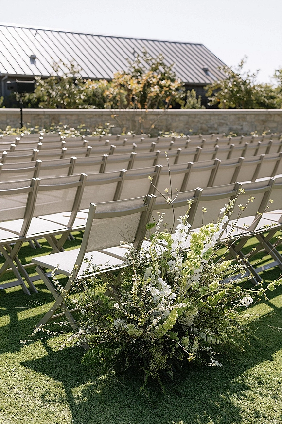 Ceremony aisle decor with aisle marker flowers and greenery along chair rows, with low floral ground arrangement on a lawn near stone wall
