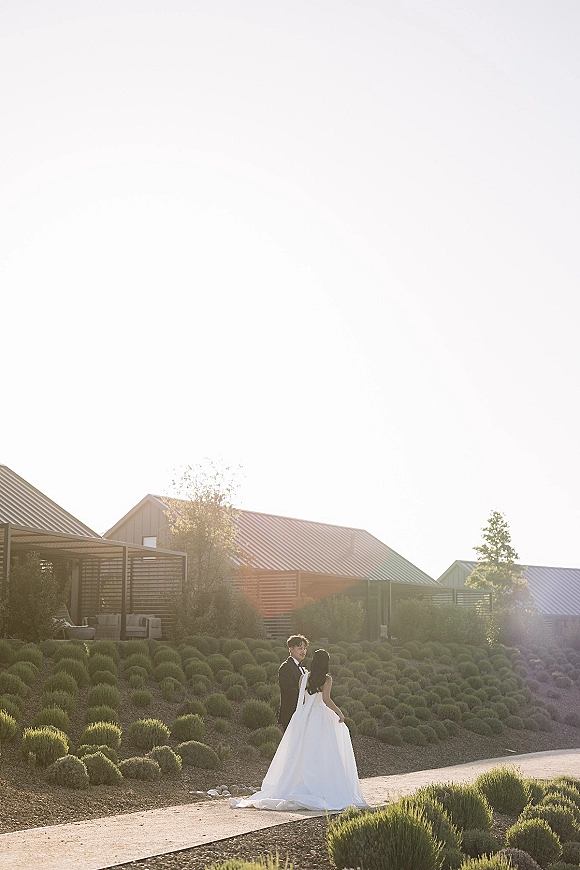 Couple portrait of bride and groom outdoors, bride holding her long train and veil as they embrace on a gravel path at sunset.