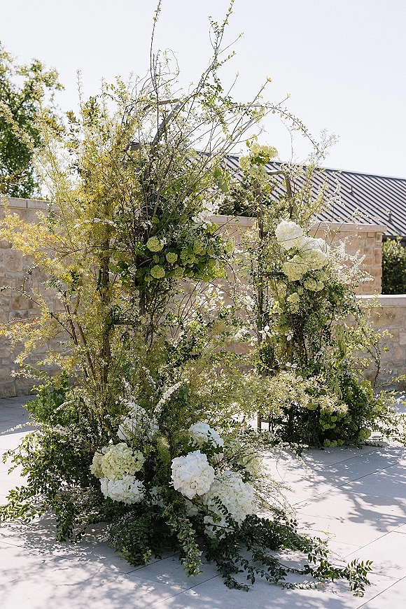 Wedding ceremony florals in a ceremony floral installation with white hydrangeas, greenery, and branches against a stone wall rooftop patio