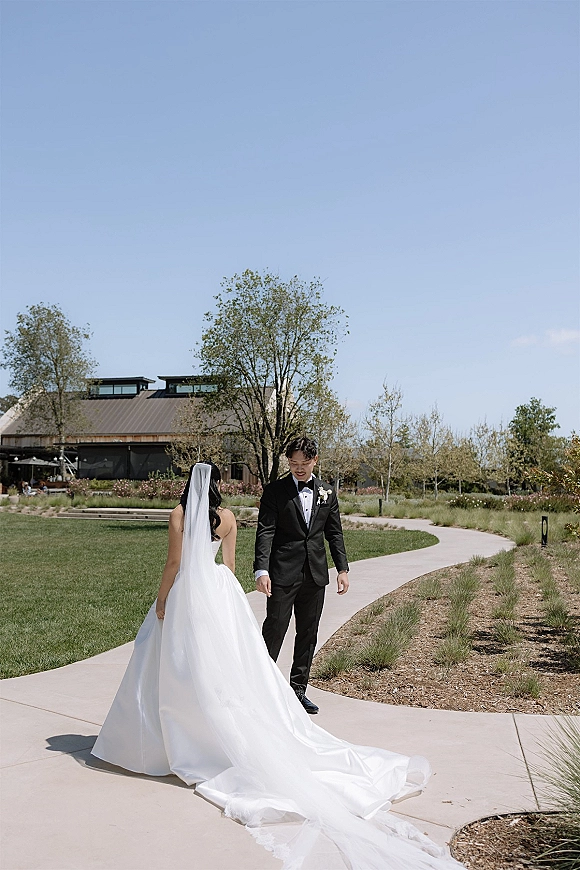 First look moment as bride in a long train wedding dress and veil walks to groom in black tuxedo on a curved garden walkway under blue sky