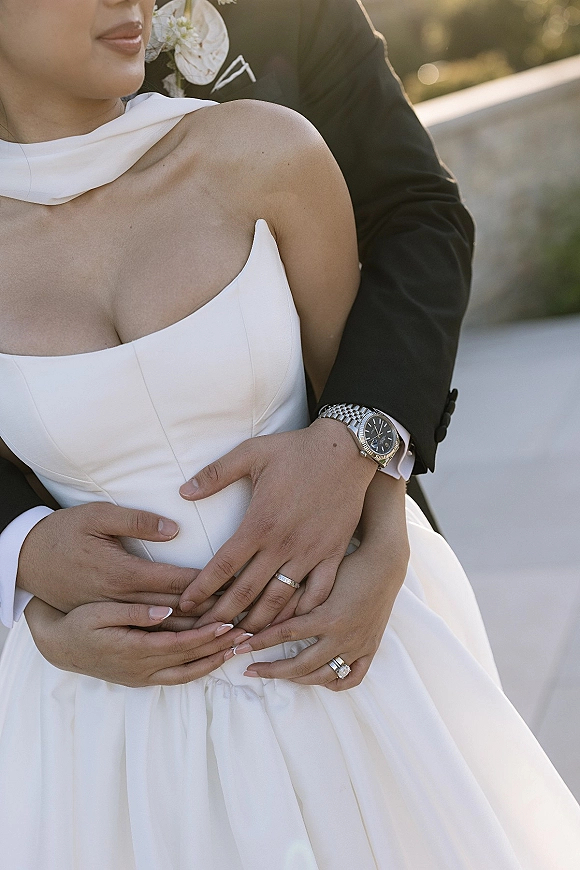 Couple portrait of bride and groom embrace, holding hands with wedding rings beside a sunlit stone railing and greenery backdrop