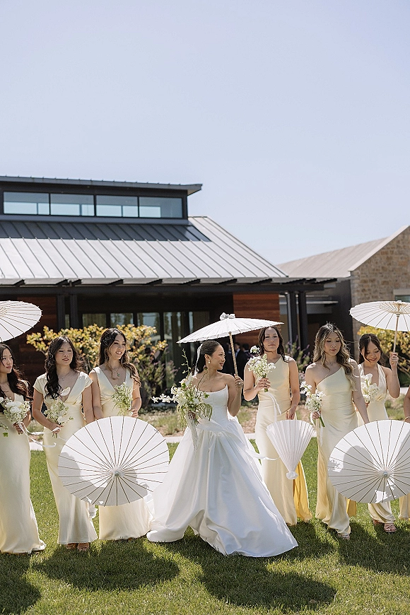 Bride with bridesmaids walking together holding white bouquets and paper parasols with ribbon streamers on a lawn by a modern venue