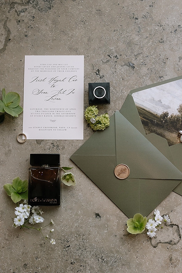 Invitation suite wedding invitation flat lay with olive green envelopes, wax seal, rings, perfume, and flowers on a stone surface