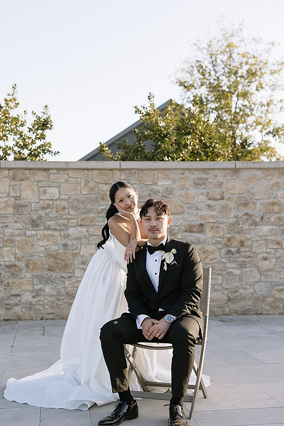 Couple portrait of bride in a strapless wedding dress standing behind groom in black tuxedo on a patio by a stone wall at golden hour