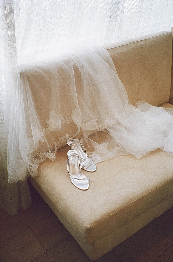 Bridal shoes, silver bridal heels with lace-trim veil draped over strappy sandals on a beige sofa in soft window light
