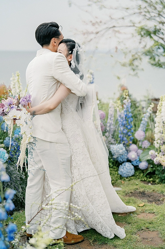 Couple portrait of bride and groom hug, her lace dress and long veil beside pastel bouquet on a coastal garden lawn with ocean backdrop