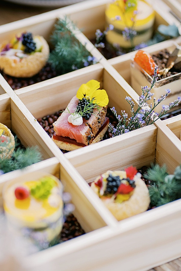 Wedding appetizer display of bite-sized canapes in a wooden compartment tray, topped with edible flowers, berries, and herbs on a table surface