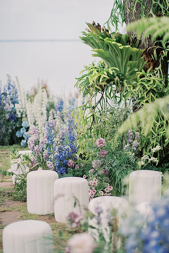 Ceremony aisle decor with pastel floral arrangements of blue delphinium, lavender blooms, and pink roses framing white ottomans by a tree trunk