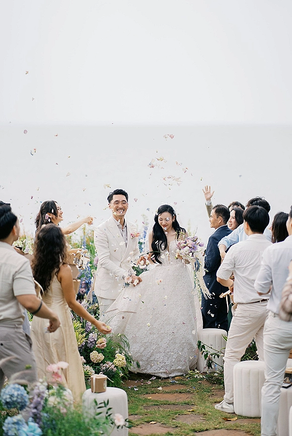 Wedding recessional with confetti wedding exit as newlyweds walk down an outdoor aisle, bride holding bouquet while guests cheer under open sky