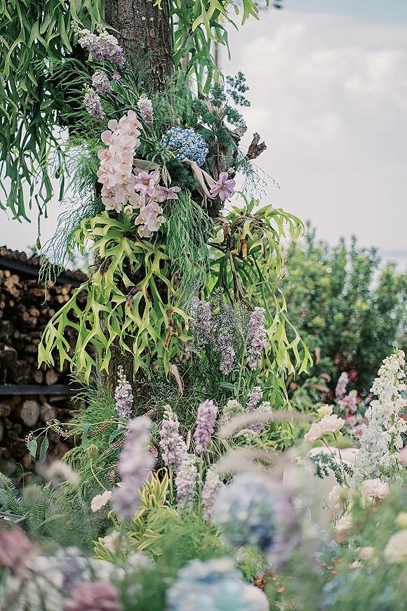 Wedding floral installation with orchids and hydrangea cascading over a tree trunk, lush greenery and ribbons in a garden setting