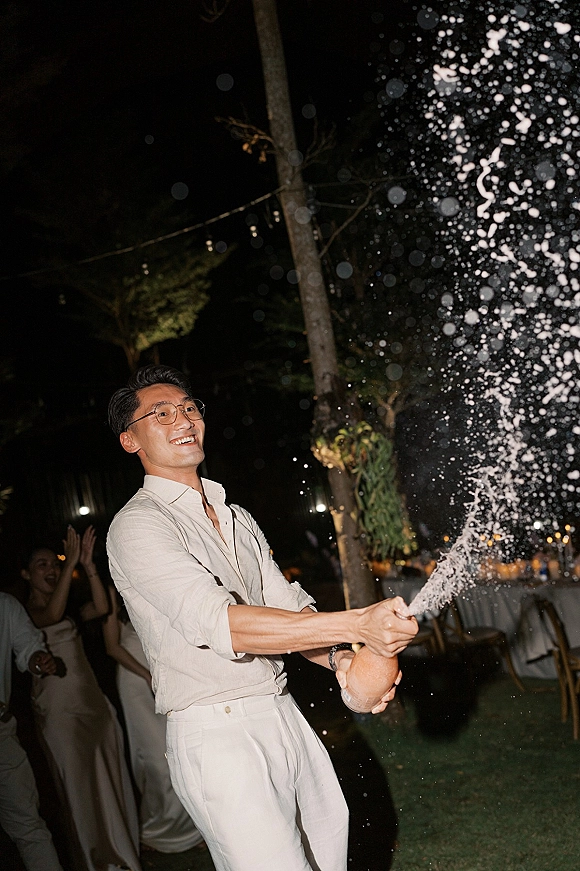 Champagne spray moment as groom pops a bottle in button-down and trousers under string lights, guests cheering at night lawn reception