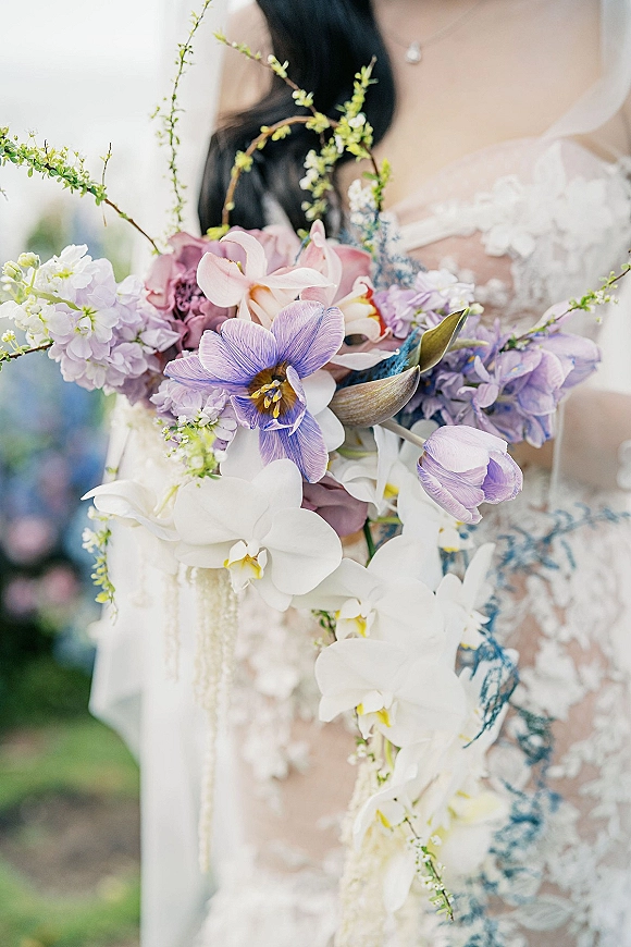 Bridal bouquet with a cascading orchid bouquet of white orchids, tulips and hydrangea held against a lace wedding dress in a blurred garden backdrop