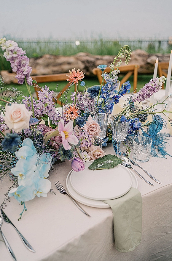 Reception tablescape with wildflower wedding centerpiece, pastel florals and taper candle, set on an ivory cloth by a stone wall and water view