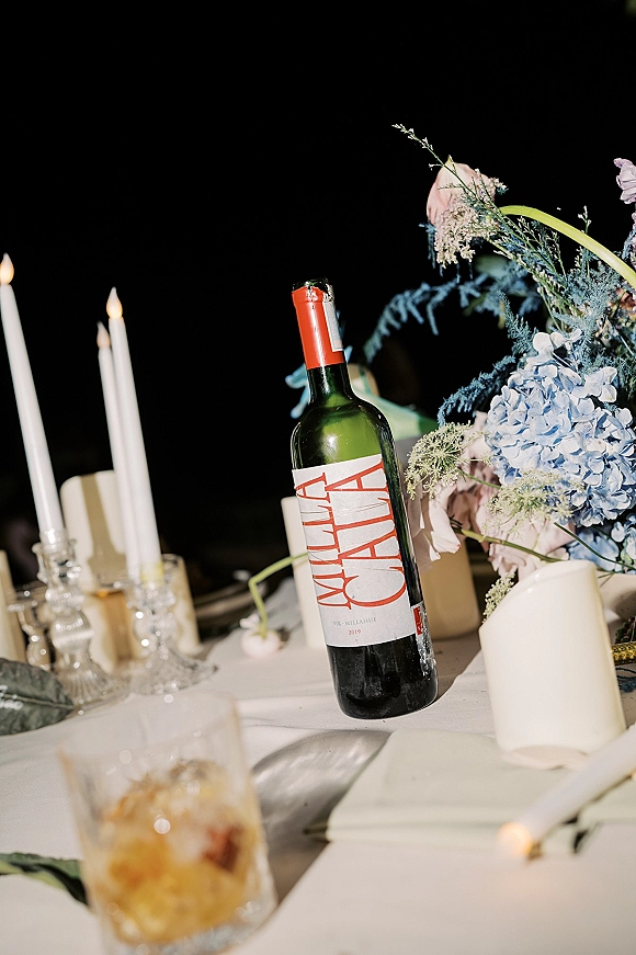 Wedding tablescape with wine bottle and taper candles beside hydrangea and rose centerpiece, glass candlesticks and menu card under night sky