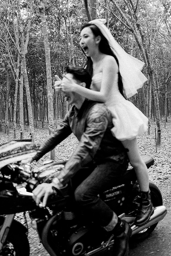 Couple portrait of bride in veil on a motorcycle behind the groom, on a leaf-strewn forest dirt road with tall trees