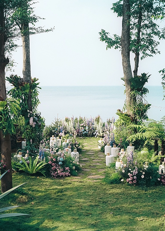Ceremony aisle design with a stone path bordered by garden florals and white lanterns, beneath trees with an ocean view beyond.