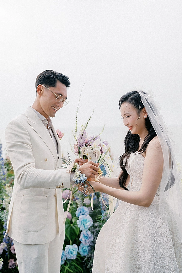 Ceremony moment as couple exchanging vows, holding hands beneath a floral arch in an outdoor garden, bride in lace dress and veil