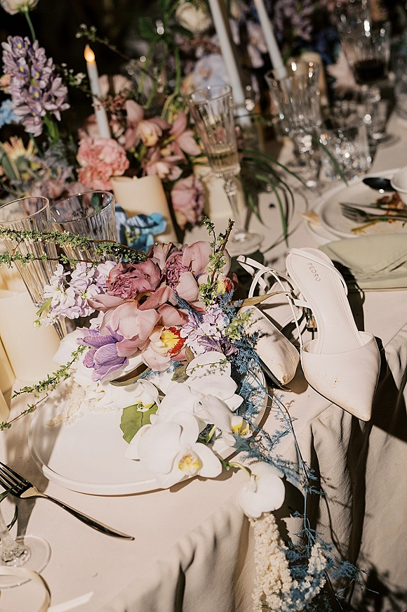 Wedding tablescape with floral centerpiece and taper candles, featuring reception table decor in dim ambient lighting with bridal heels beside glassware