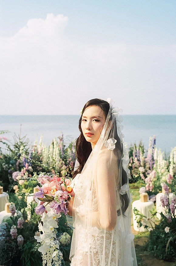 Bridal portrait of a bride holding bouquet, veil over face in a strapless lace dress with cascading orchids against an ocean horizon backdrop