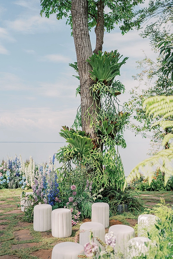 Outdoor wedding decor with tree floral installation, hanging greenery and staghorn ferns around white pedestal stands on a lawn by water