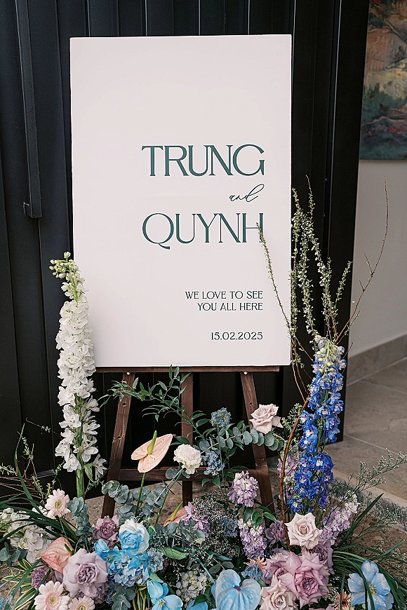Wedding welcome sign on an easel, acrylic wedding welcome sign framed by roses, delphinium, and eucalyptus against a black paneled wall