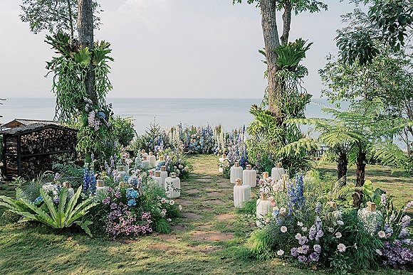 Ceremony aisle decor with pastel florals and white lanterns lining a stone path, hanging greenery in trees, and ocean view beyond