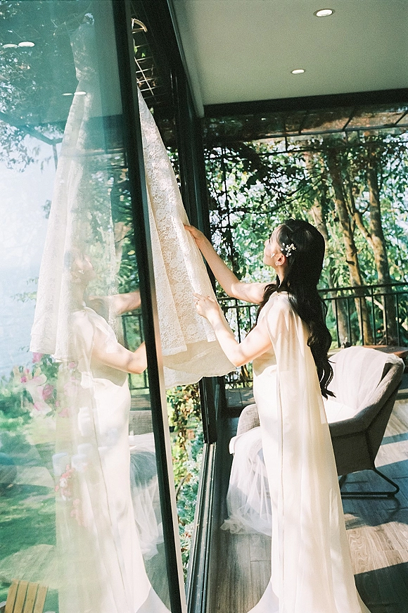 Bridal getting ready as the bride hanging wedding dress by window, wearing a lace robe with pearl hair pins in soft daylight by balcony greenery