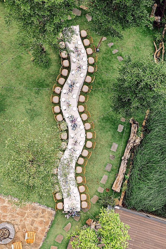 Outdoor reception tablescape with a long banquet table, white linens, floral garland and candles on a garden lawn under string lights