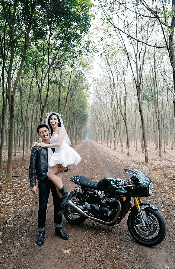 Couple portrait of bride on groom’s piggyback pose, laughing in a veil and leather jacket beside a motorcycle on a forest dirt road
