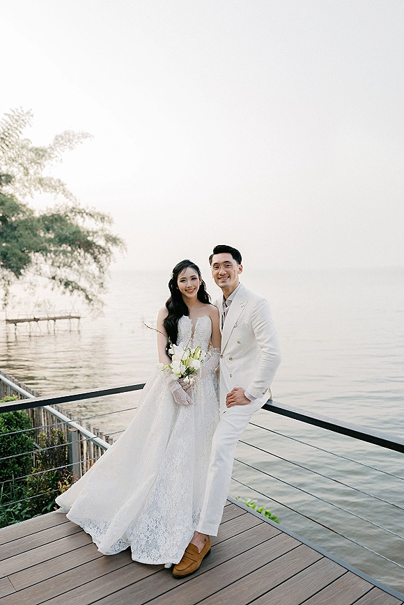 Couple portrait of bride in lace dress and veil holding a white bouquet beside groom in white suit on a lakeside dock railing