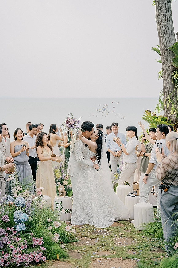 Wedding kiss at an ocean view wedding ceremony, bride in lace dress with veil and bouquet as guests toss flower petals along aisle florals