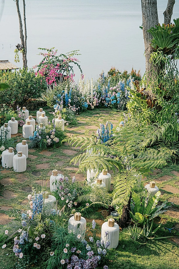 Ceremony aisle decor with ground florals and white lanterns lining a stone path, framed by lush garden greenery with water view
