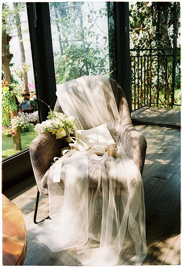 Wedding details with bridal veil draping over an armchair beside a bouquet, invitation suite, ribbon and ring box by sunlit windows overlooking trees