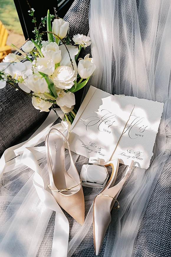 Wedding flat lay of bridal details with invitation, vow books, perfume, shoes and bouquet arranged on an upholstered chair in window light