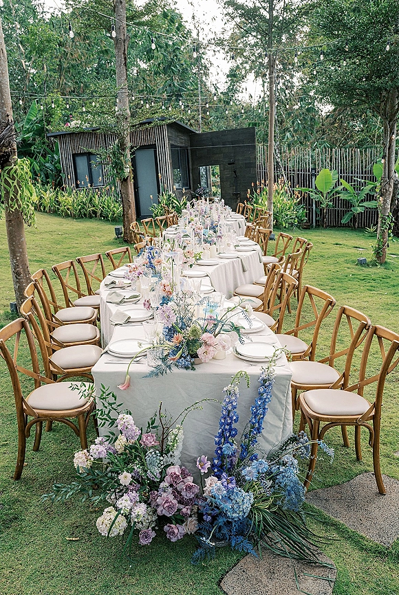 Reception tablescape with a long banquet table, white linens, pastel centerpieces, and string lights on a garden lawn by a modern building