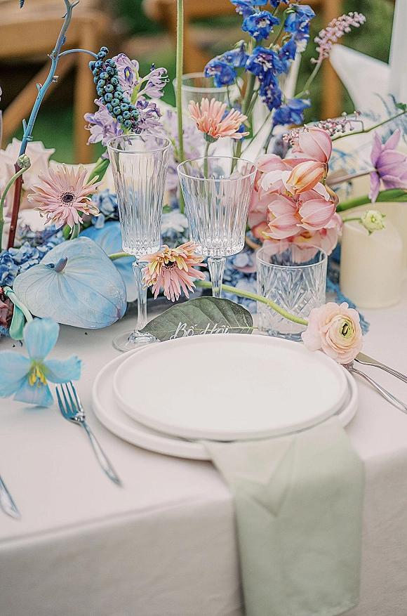 Reception tablescape with wedding table setting, bud vase of pastel blooms beside white plates, sage green napkin, and clear stemware on white linen