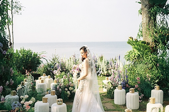 Bridal portrait of a bride holding bouquet with a long veil, lace wedding dress, and floral hair piece in an ocean-view garden setting