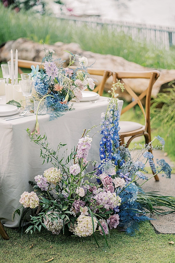Reception tablescape with an outdoor wedding tablescape featuring pastel floral centerpiece and taper candles on a white tablecloth on a lawn with rocks