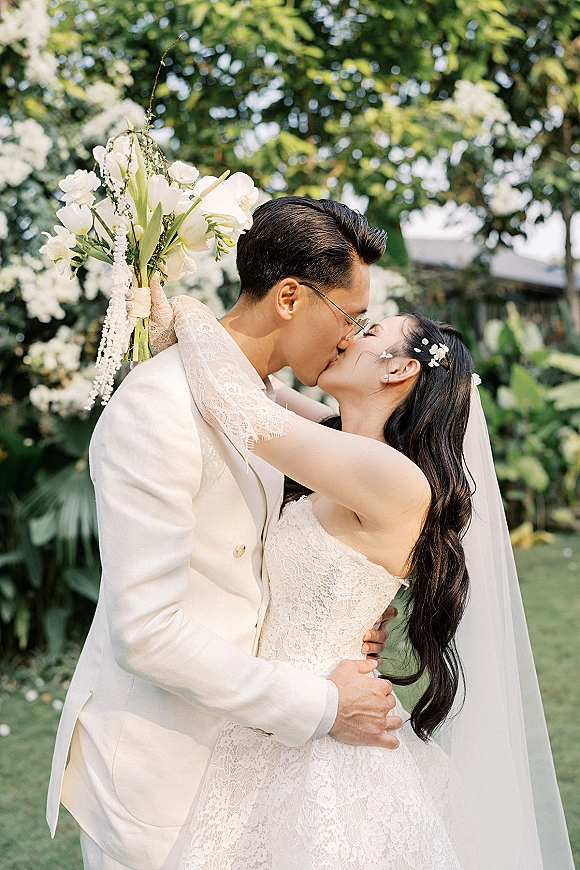 Wedding kiss as bride and groom kissing in a lush garden, her veil and lace dress flowing while she holds a white bouquet