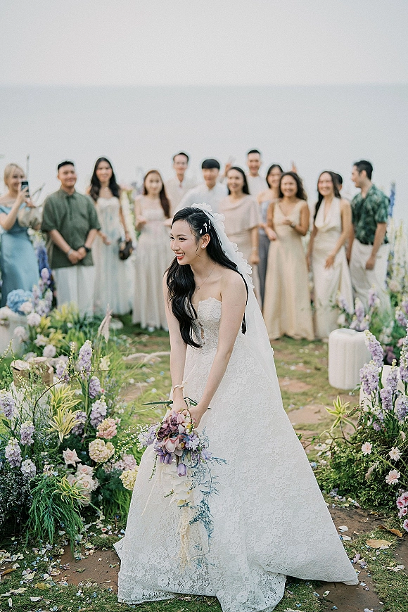 Bouquet toss moment as bride in strapless lace dress and veil throws cascading bouquet on outdoor lawn with guests behind under sky