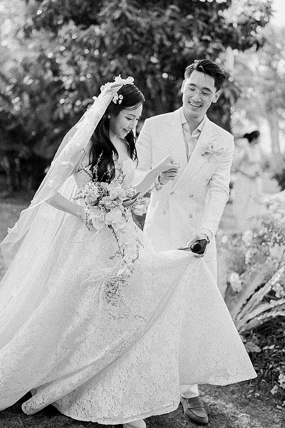 Wedding couple portrait of bride and groom walking as he holds a champagne bottle, her veil and bouquet flowing on a garden walkway