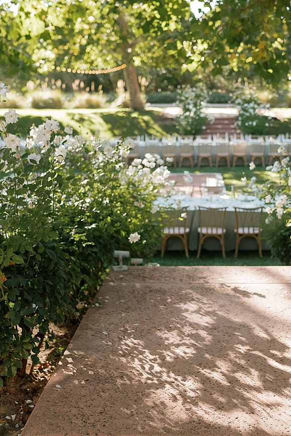 Outdoor reception setup with long banquet tables, wooden chairs, sage linens, and string lights on a dappled garden lawn by stone steps