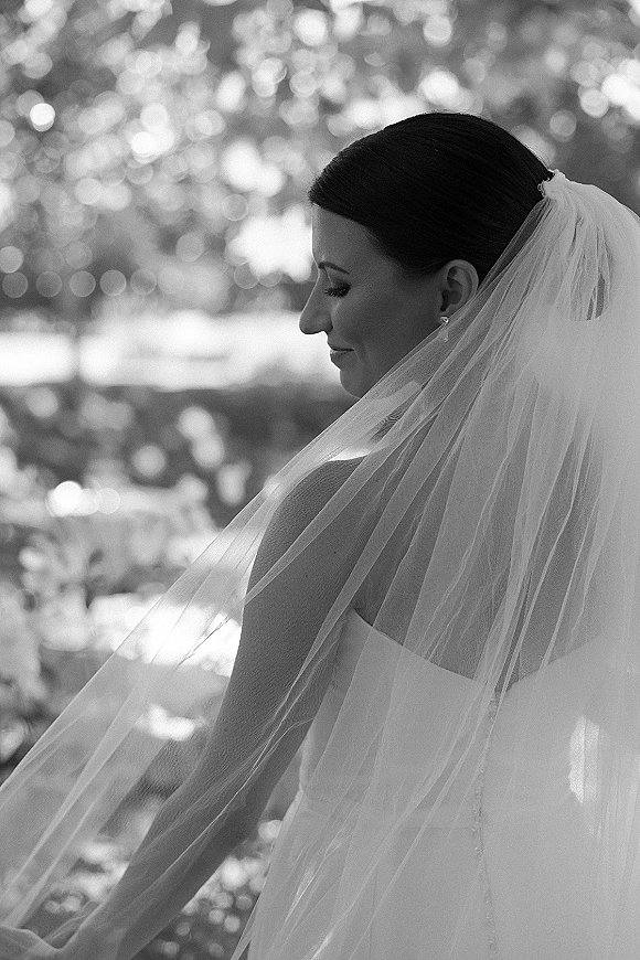 Bridal portrait in black and white of a bride in veil, wearing a strapless dress and drop earrings, with sunlit garden bokeh behind